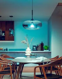 Hanging pendant lamp over a dining table, set to a deep blue Hue color, casting teal light across the table, the chairs, and the kitchen behind.