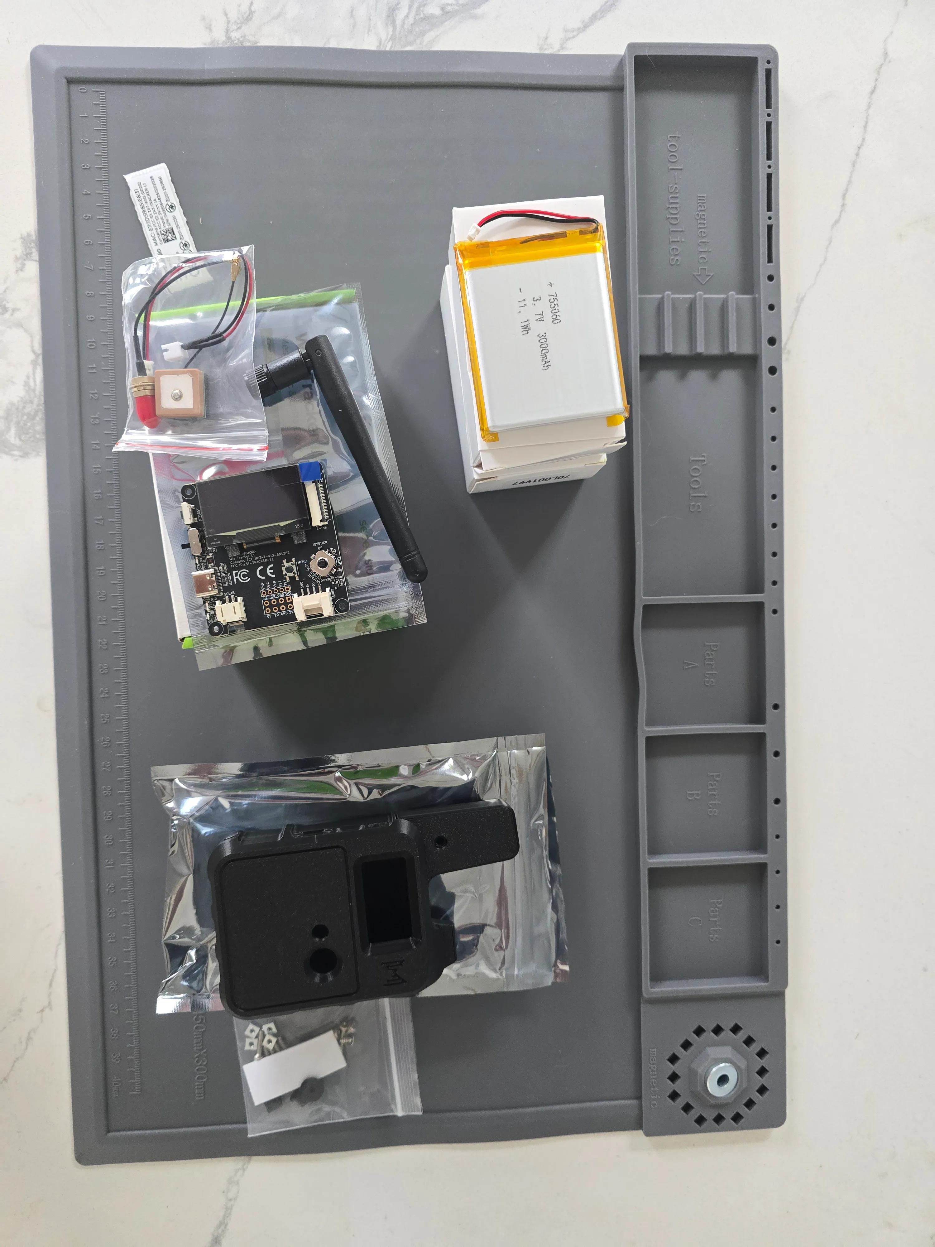 Wio Tracker L1 unboxed on a work mat: a 3.7-volt 3000 mAh lithium pouch cell, the Wio-SX1262 board with a stubby LoRa antenna and a GPS patch antenna on a u.FL lead, a 3D-printed handheld enclosure, and a bag of assembly screws.